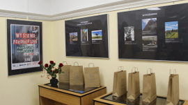 fotografia przedstawiająca wystawę prac nagrodzonych w konkursie, wyeksponowanych w ramach oraz torby papierowe z nagrodami i róże w wazonieWystawa prac nagrodzonych